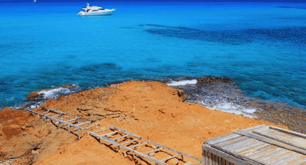 "A scenic view of Cala Saona beach with a wooden ramp leading down to the rocky shore, crystal-clear turquoise waters, and a yacht anchored in the distance."