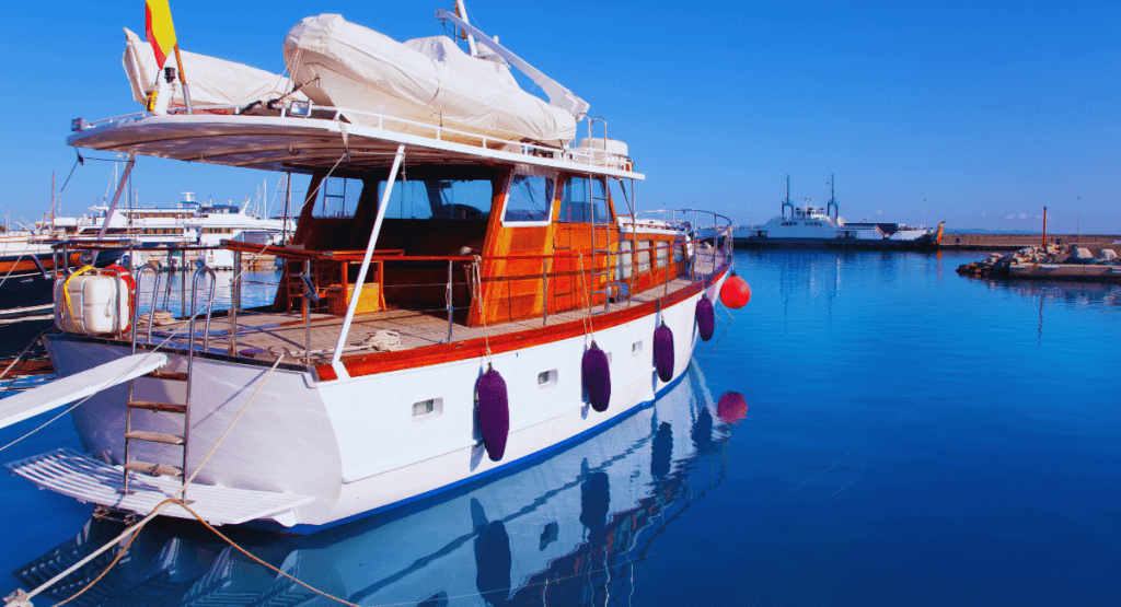 "A wooden yacht docked at La Savina Port, surrounded by calm blue waters and other boats in the background under a clear blue sky."