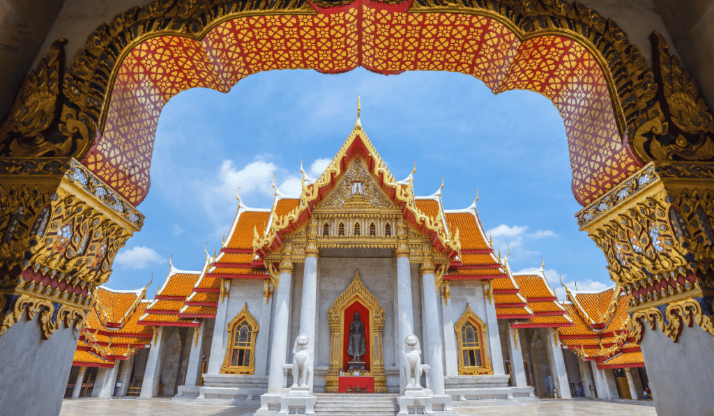 The Marble Temple (Wat Benchamabophit), a beautiful marble temple in Bangkok, Thailand. The temple has a red roof, gold details, and white columns. There are two large lion statues guarding the entrance. The sky is blue with white clouds.