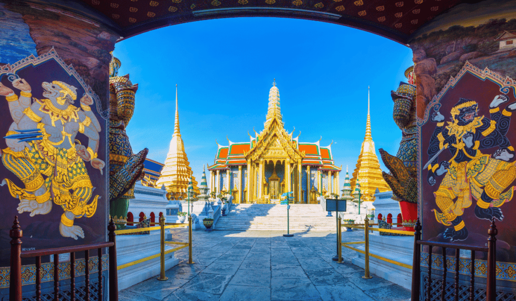 A grand and ornate temple complex in Bangkok, Thailand. The main building is a towering structure with golden spires and intricate details. Two large, mythical creatures stand on either side of the entrance, guarding the temple. The sky is blue and clear.