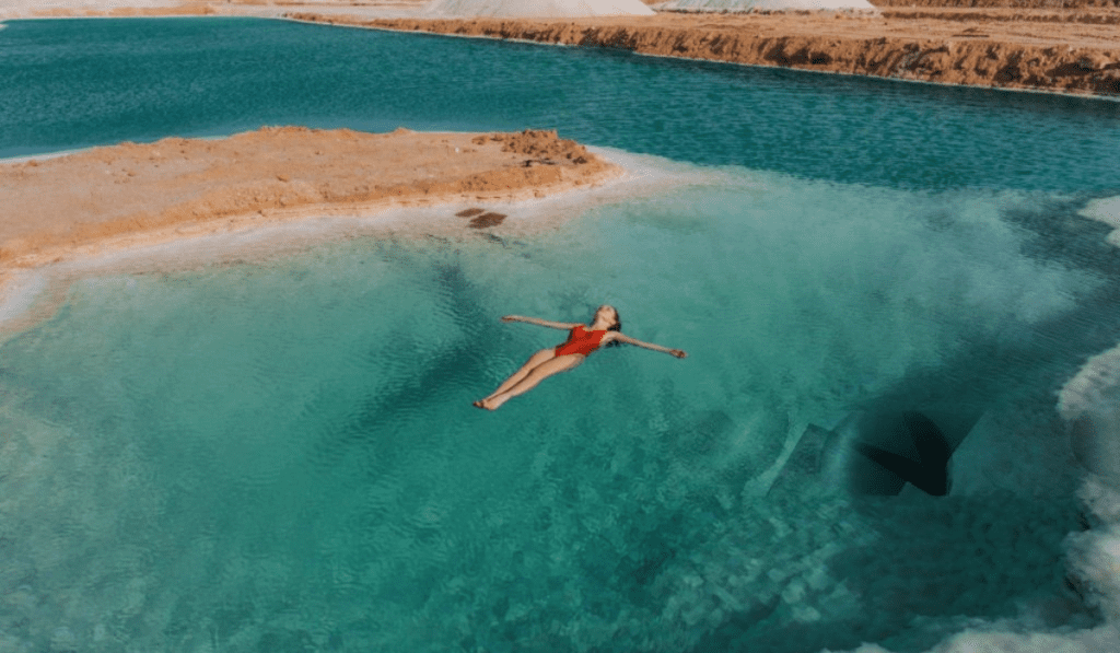 Best Time to Visit Siwa Salt Lake egypt A woman in a red swimsuit is floating on her back in a small, turquoise Siwa Salt Lake egypt surrounded by salt-crusted cliffs. The water is clear and calm, and the sky is a bright blue.