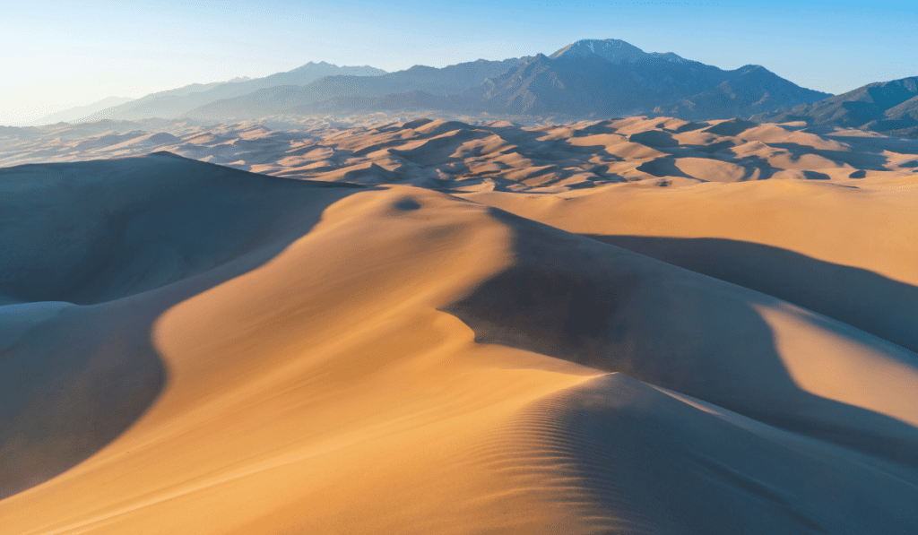 Great Sand Sea Great Sand Sea, a vast expanse of sand dunes stretching towards a distant mountain range. The dunes are golden brown and cast long shadows in the early morning light.