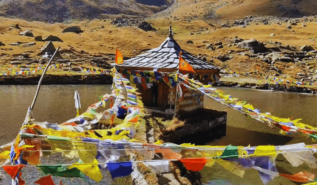 The Yulla Kanda Temple, a small, wooden temple with a conical roof, sits on a platform in the middle of a serene lake, surrounded by colorful prayer flags. The lake is nestled in a mountainous landscape with rolling hills and rocky terrain.