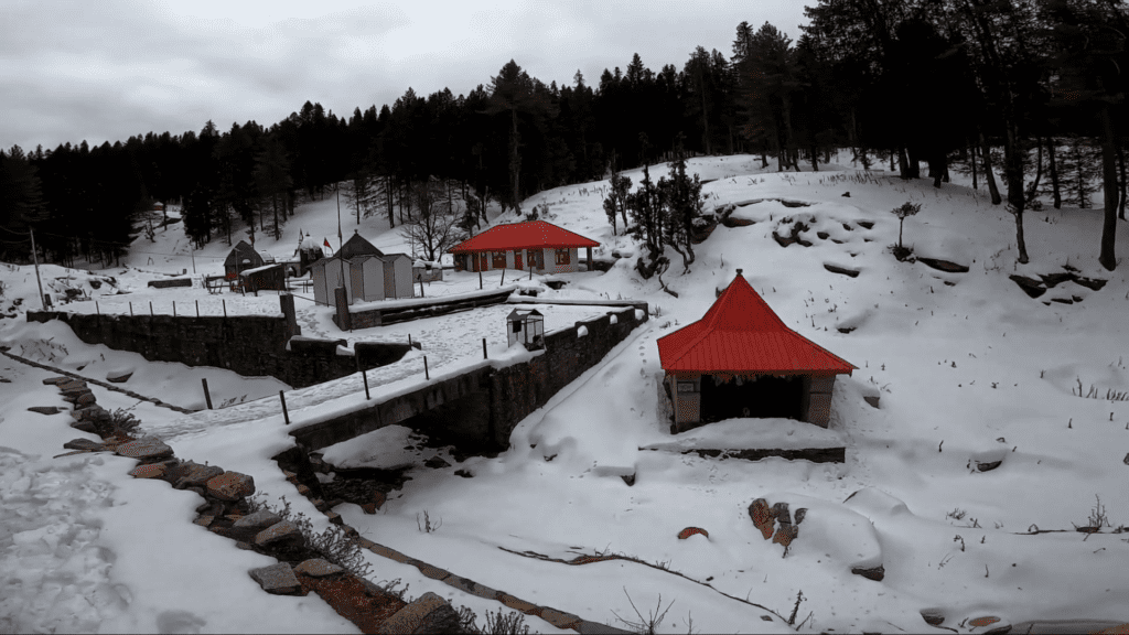 Giri Ganga Temple, a serene Hindu shrine blanketed in snow, located in Giri Ganga, Himachal Pradesh. The temple complex features a main shrine with a red roof, connected to a smaller shrine by a stone bridge. Surrounding the complex are snow-covered hills and pine trees.