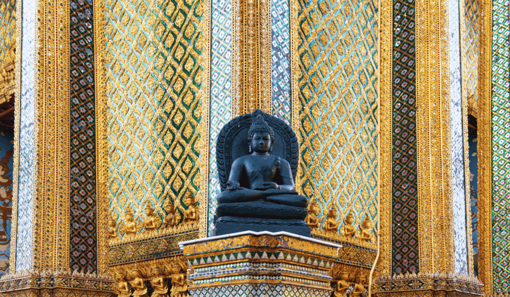 A black stone Buddha statue seated in a meditative pose, set against an ornate golden and green patterned wall at Wat Phra Kaew in Bangkok, Thailand."