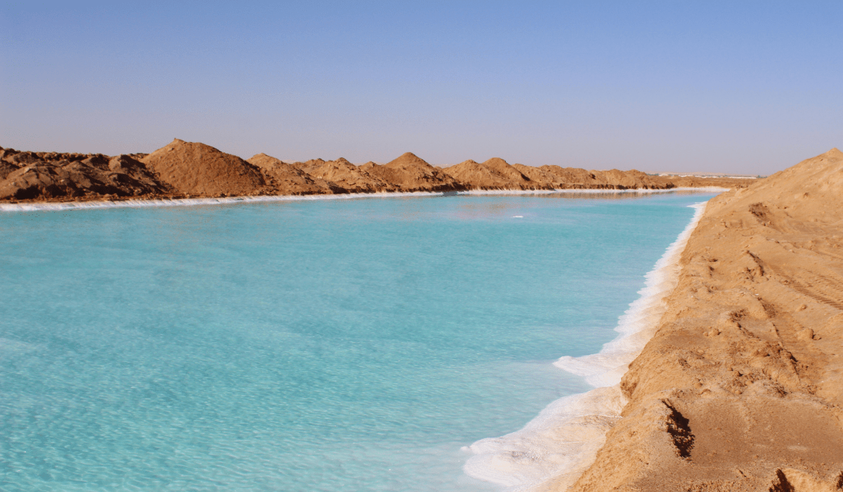 A clear blue salt lake surrounded by sandy terrain in Siwa Salt Lake Egypt, with visible salt deposits along the water's edge, under a bright and clear sky.