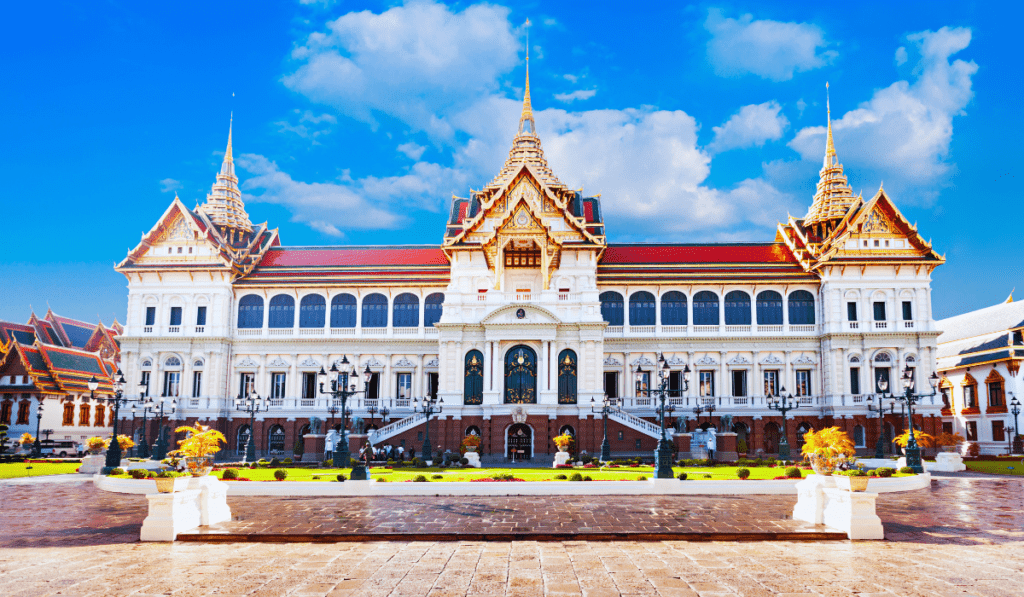 A grand and ornate palace building in Bangkok, Thailand. The building is white with gold details and has a red roof. There are tall spires on either side of the building. The sky is blue with white clouds.