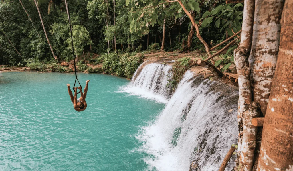Things to Do in Cambugahay Falls A visitor enjoying a thrilling rope swing over the turquoise waters of Cambugahay Falls in Siquijor, Philippines. The person is mid-air, having just launched off the swing, with the waterfall cascading in the background and surrounded by lush greenery. The scene captures the adventurous spirit and natural beauty of Cambugahay Falls, a popular activity for tourists seeking excitement and fun.