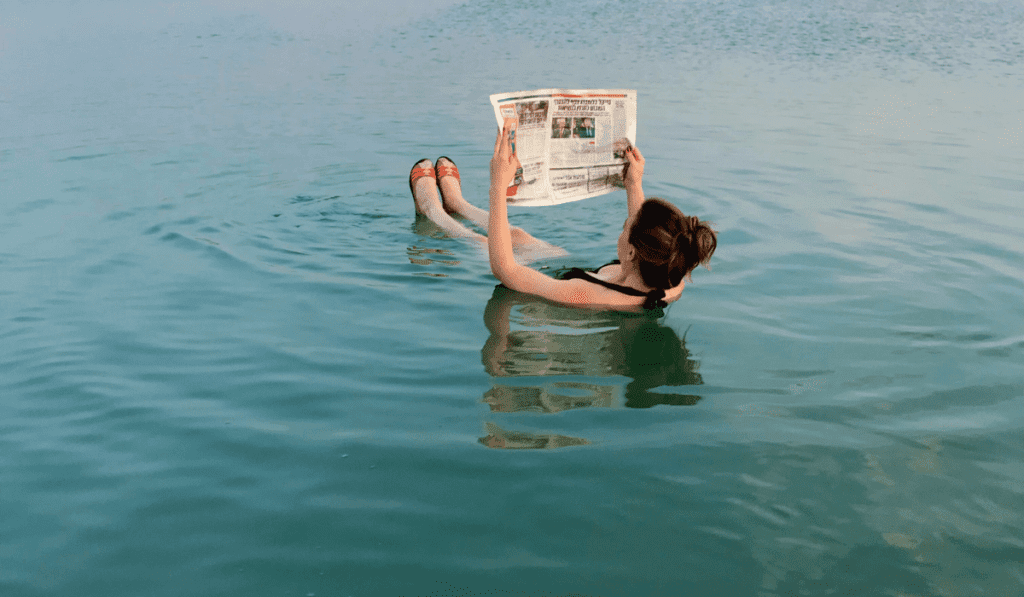 Things to Do in Siwa Salt Lake A woman leisurely floats in the Siwa Salt Lake egypt, engrossed in reading a newspaper, surrounded by a serene aquatic environment.