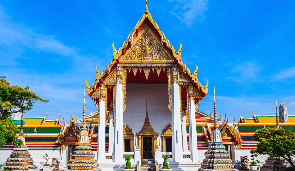 Wat Pho, a famous temple in Bangkok, Thailand, featuring a red roof, gold details, and white columns. The temple is surrounded by smaller structures and lush greenery. The sky is blue with white clouds.