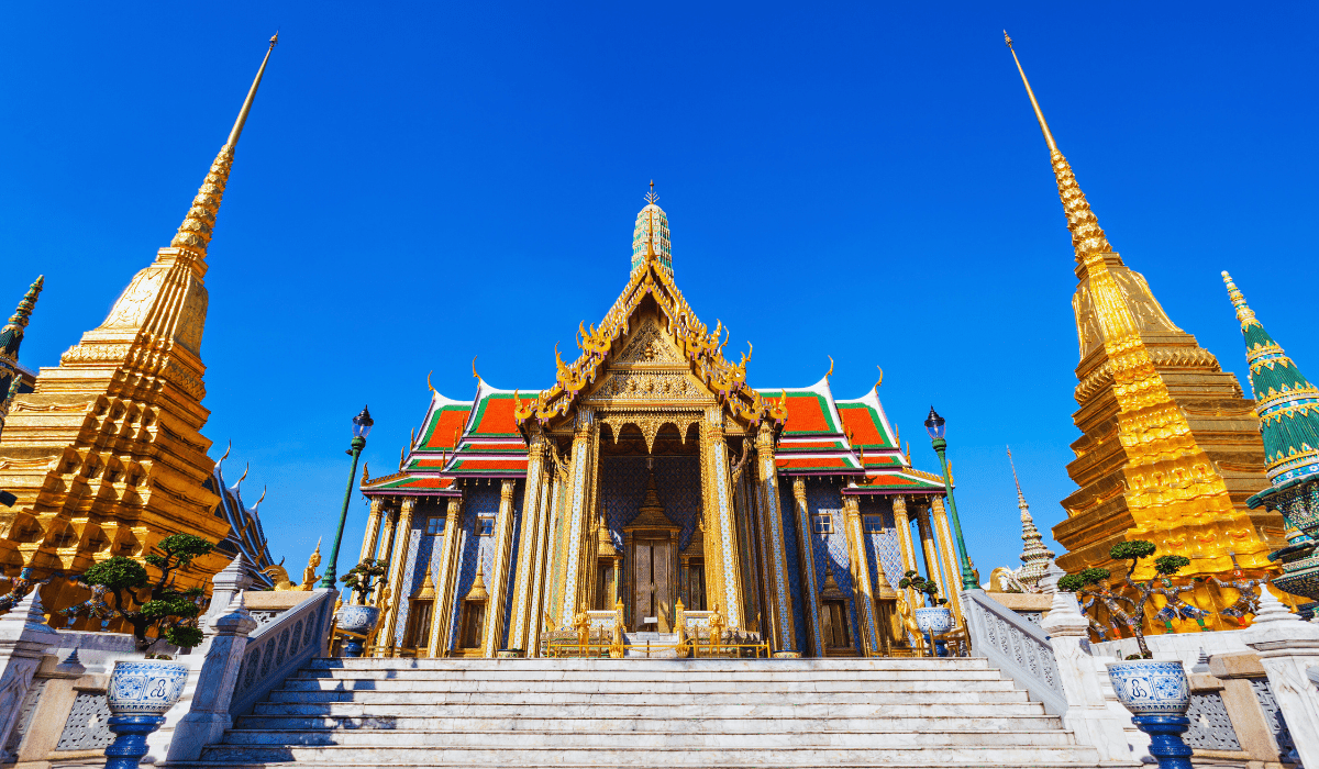 "The grand Wat Phra Kaew temple with golden spires and intricate architectural details, located in Bangkok, Thailand, under a clear blue sky."