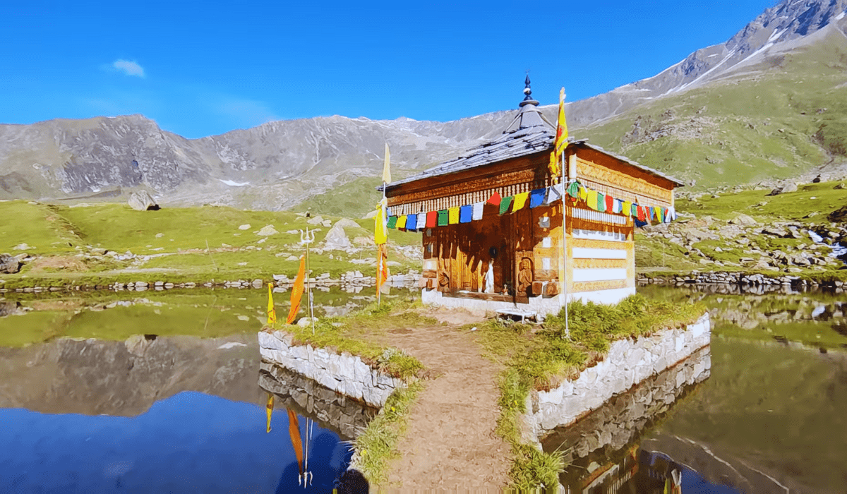 : A small wooden temple stands on an island surrounded by a calm lake at Yulla Kanda, set against a backdrop of towering green mountains. The temple, adorned with colorful prayer flags, reflects in the clear waters, enhancing the serene atmosphere of this high-altitude sacred site.