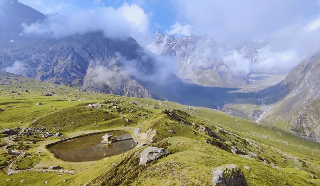 A scenic view of Yulla Kanda, featuring a small temple situated on a grassy plateau surrounded by mountains. The temple is reflected in a nearby lake, and mist rises from the valley below.