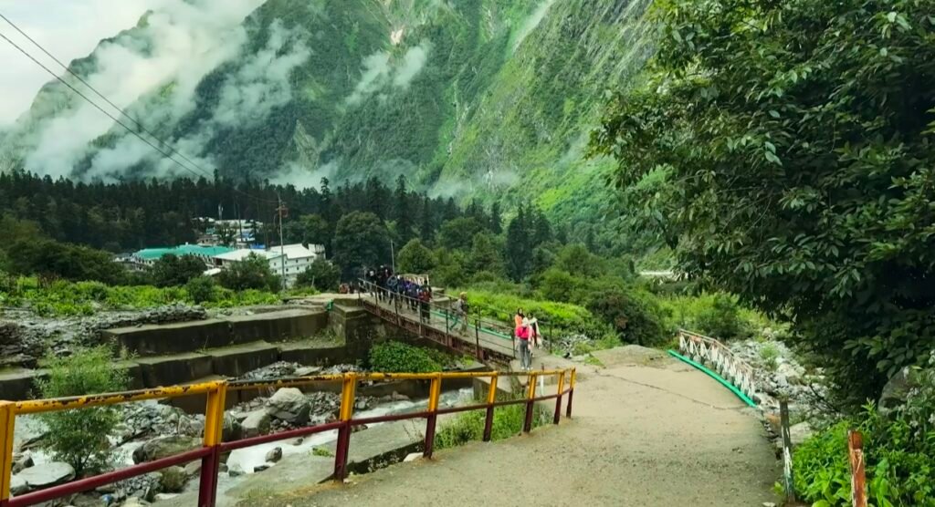 Hikers crossing a small bridge over a rocky stream, set against lush green mountains partially shrouded in clouds, with a village and pine trees nestled at the base of the valley on the Valley of Flowers trek in Uttarakhand, India.