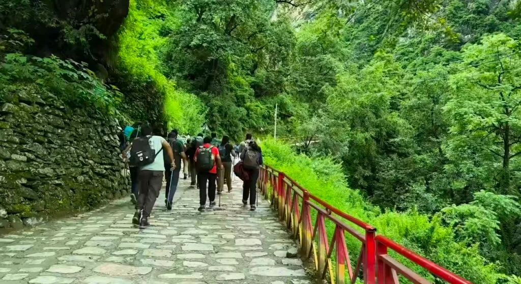 Group of trekkers walking along a paved stone pathway bordered by a red railing, surrounded by lush green forest and dense vegetation in the Valley of Flowers, Uttarakhand, India. The path winds through the foothills of the Himalayas, offering a scenic view of hillside trees, mossy stone walls, and vibrant natural greenery typical of this UNESCO World Heritage Site trek route