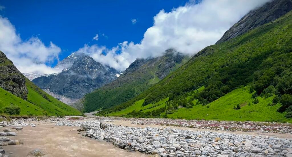 Snow-capped Himalayan peak rising above a lush green valley, with steep forested hillsides and a rocky riverbed under a bright blue sky and scattered white clouds, typical of the Valley of Flowers trek in Uttarakhand, India.