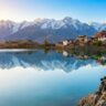 A backpacker sits on rocky terrain overlooking the serene Nako Lake in Kinnaur, Himachal Pradesh, with snow-capped Himalayan mountains reflected in the calm water. Traditional village houses and a monastery are visible along the lake shore under a clear blue sky, illustrating the tranquil beauty and cultural charm of this high-altitude Himalayan destination.