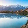 A backpacker sits on rocky terrain overlooking the serene Nako Lake in Kinnaur, Himachal Pradesh, with snow-capped Himalayan mountains reflected in the calm water. Traditional village houses and a monastery are visible along the lake shore under a clear blue sky, illustrating the tranquil beauty and cultural charm of this high-altitude Himalayan destination.