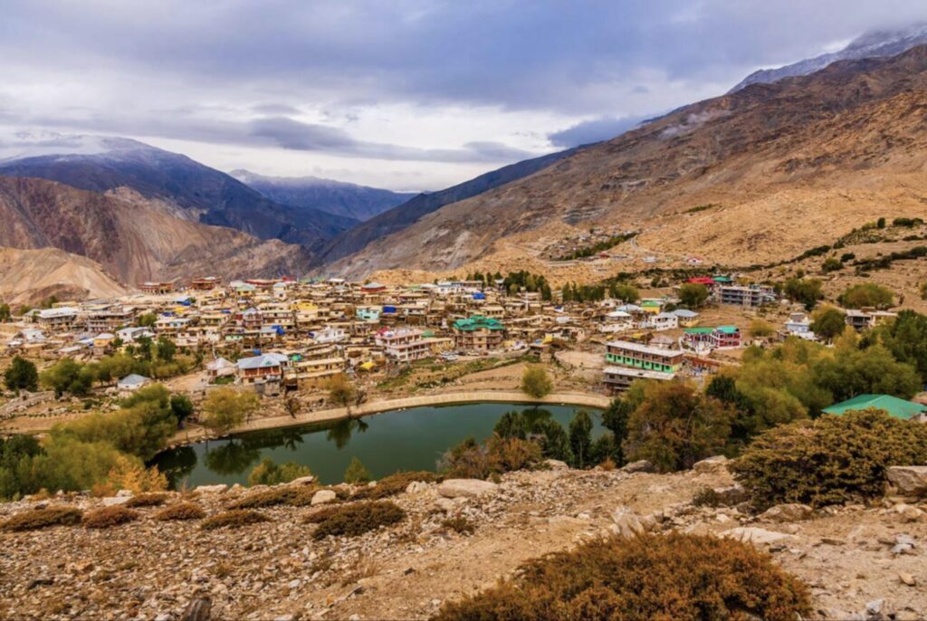 A scenic view of Nako Village in Kinnaur, Himachal Pradesh, India, nestled in a high-altitude Himalayan valley. The picturesque village with colorful flat-roofed houses is clustered around the serene, emerald-green Nako Lake, surrounded by golden autumn trees and barren, rugged mountains under a dramatic cloudy sky."