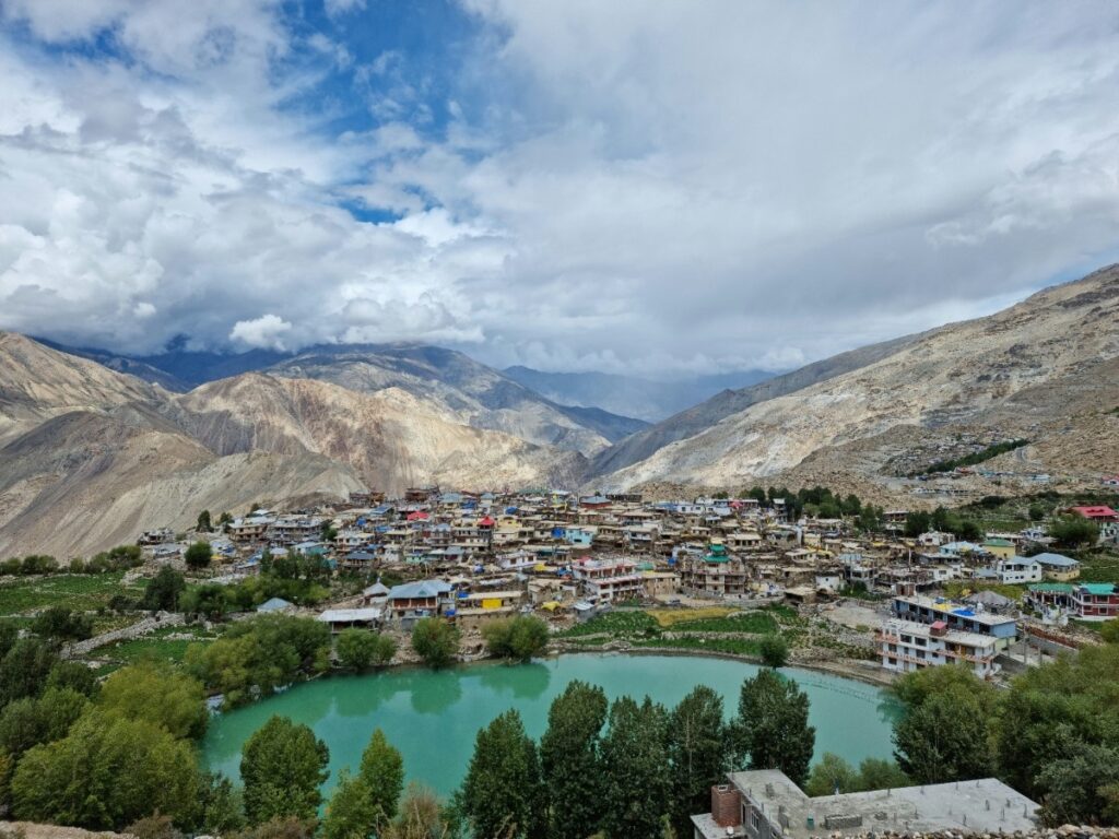 A high-angle, wide landscape photograph shows a town with many small buildings nestled in a valley above a turquoise nako lake, surrounded by arid, brown mountains under a partly cloudy sky.