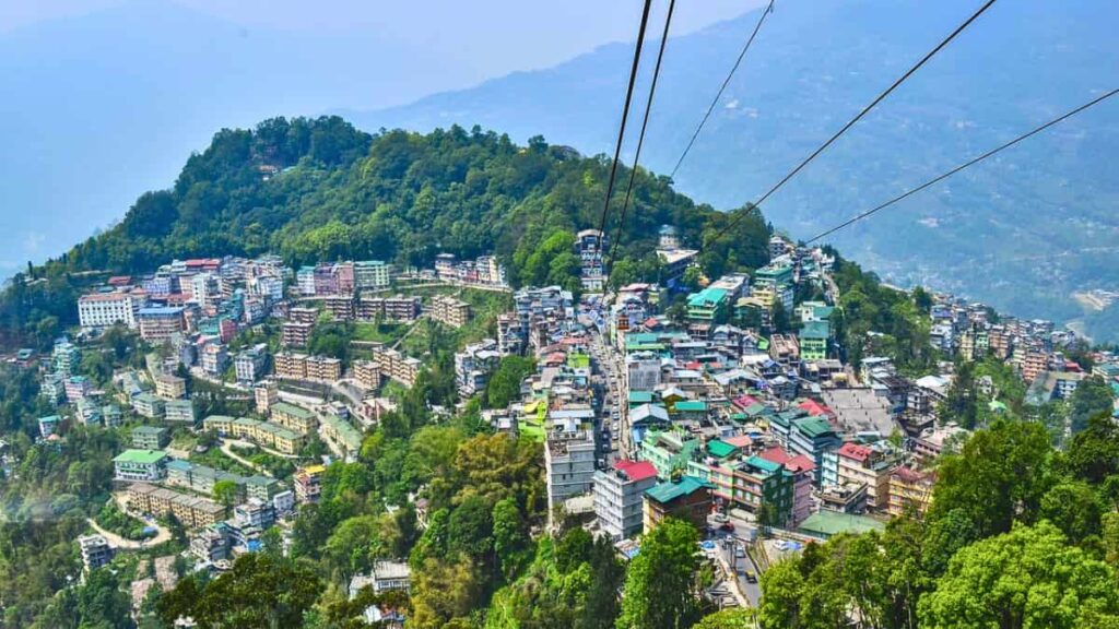 "Panoramic vista of the colorful buildings and steep hills of Gangtok, Sikkim, as seen through the cables of the city's famous aerial tramway."