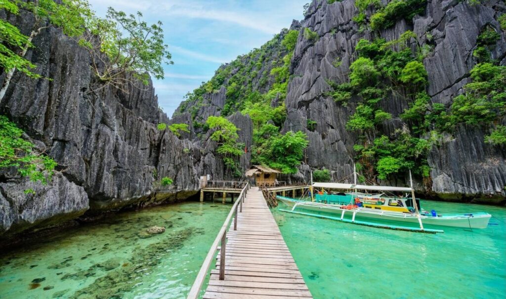 Scenic view of Barracuda Lake, Coron Island, with a wooden pier, traditional boats, emerald-green water, and towering limestone cliffs.