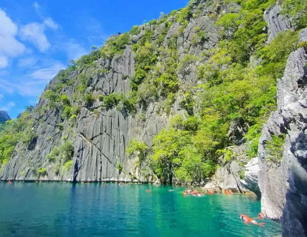 Traditional Filipino bangka boats docked in a turquoise lagoon at the entrance of Barracuda Lake, surrounded by jagged grey limestone cliffs and lush greenery in Coron, Palawan.