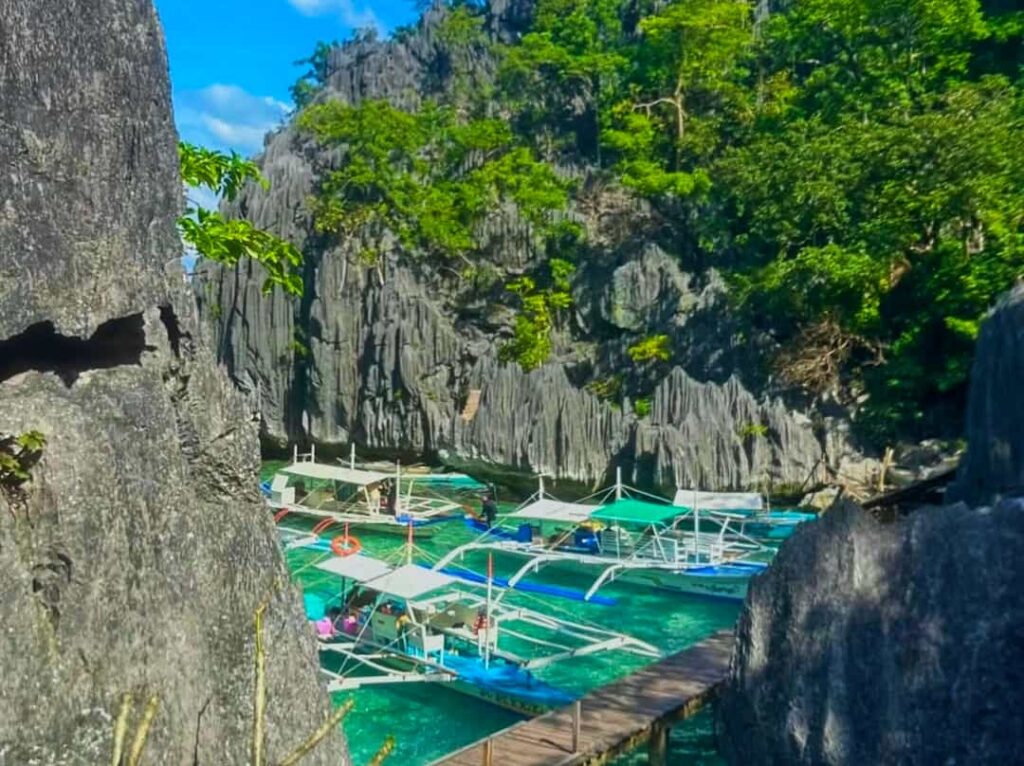 A scenic view of the entrance to Barracuda Lake in Coron, Palawan, featuring traditional bangka boats docked near a wooden walkway surrounded by dramatic limestone karst cliffs and turquoise water.