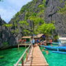 Tourists arriving at Barracuda Lake, Coron Island, via a wooden walkway with boats, crystal-clear lagoon water, and towering limestone cliffs.