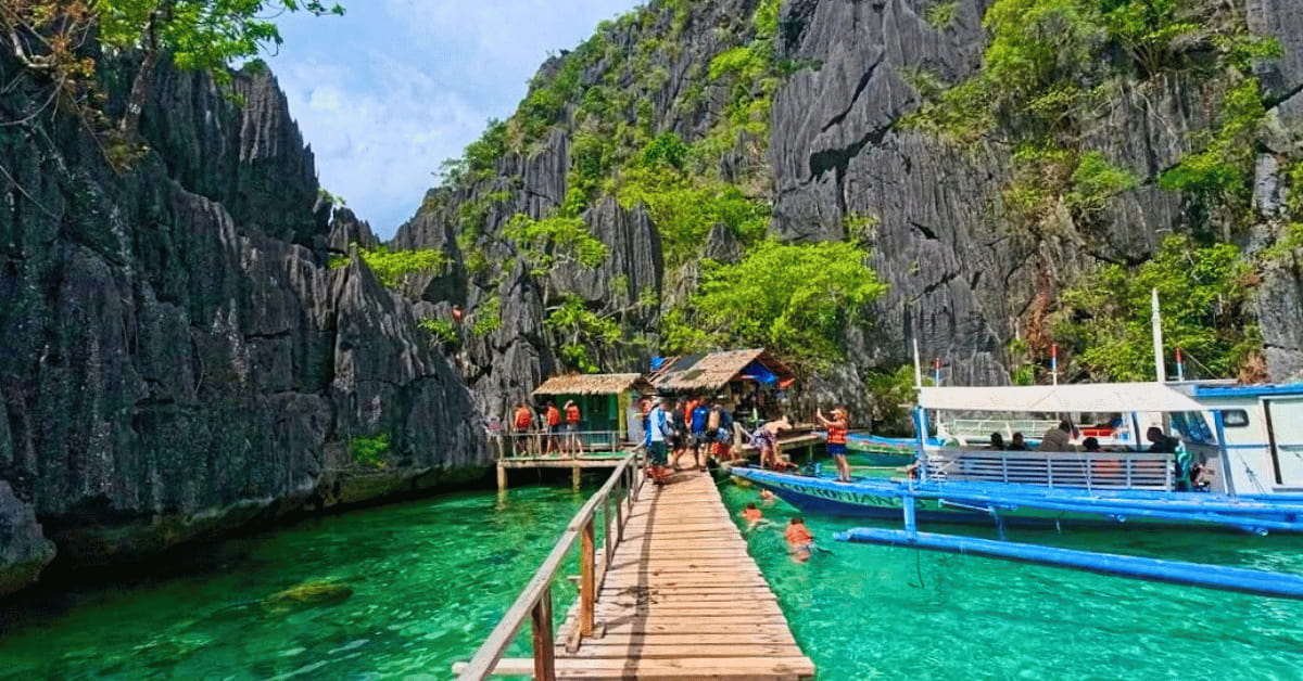 Tourists arriving at Barracuda Lake, Coron Island, via a wooden walkway with boats, crystal-clear lagoon water, and towering limestone cliffs.