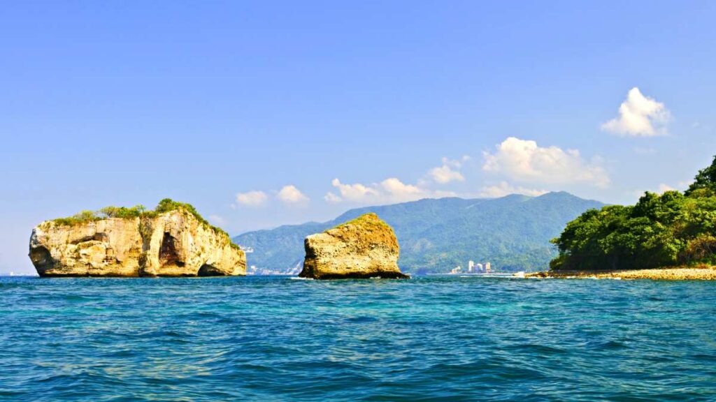 Calm ocean scene with two rocky islands covered in greenery, set against a backdrop of distant mountains and a bright blue sky with scattered clouds.
