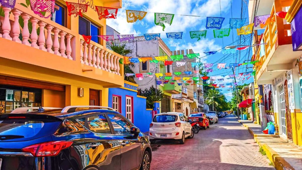 A vibrant street with colorful papel picado banners hanging above. Brightly painted buildings line the road, with parked cars and clear blue skies.